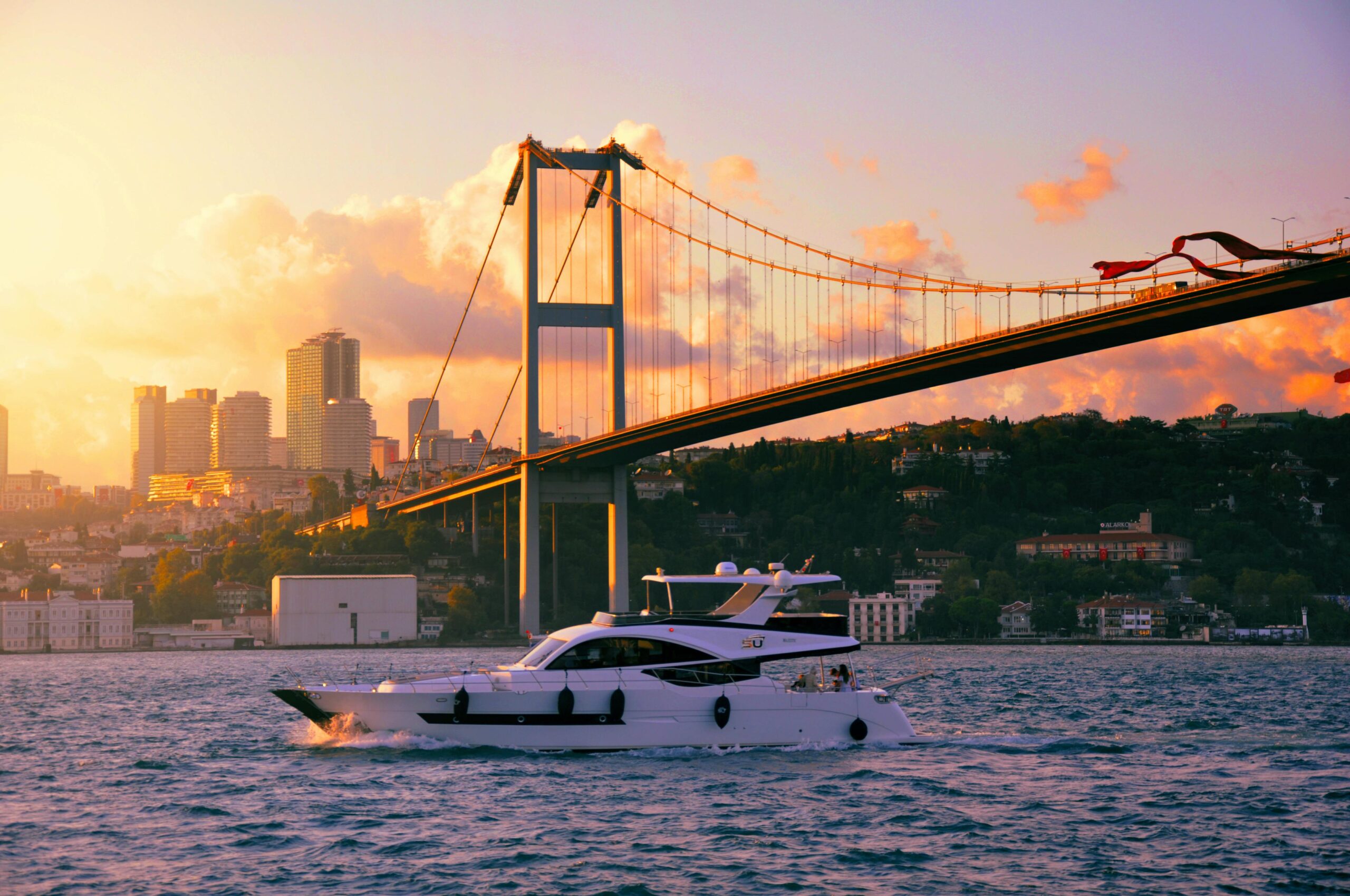 Luxurious yacht glides under the scenic Bosphorus Bridge at sunset in İstanbul, Türkiye.